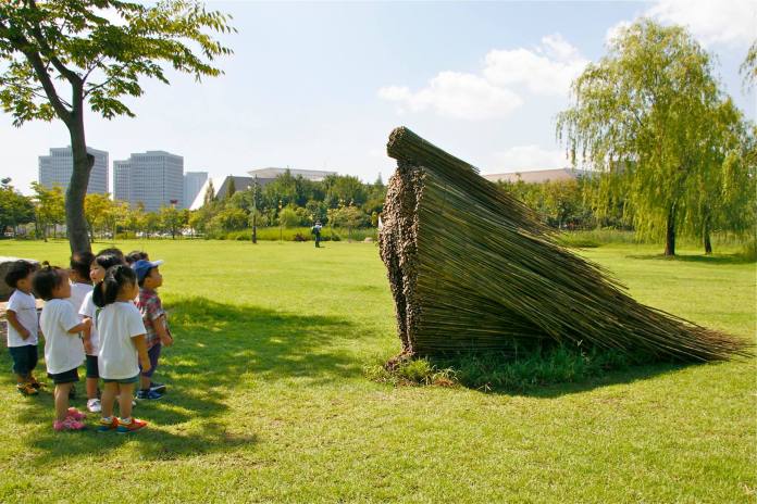Outdoor sculpture of a figure formed from bundled sticks bending forward, with children observing it on a grassy field surrounded by city buildings.