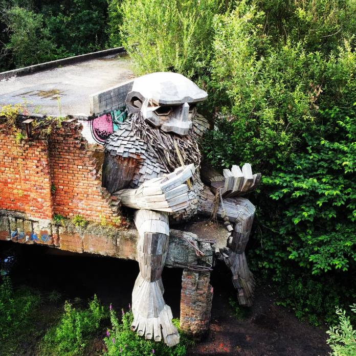 Large wooden troll sculpture with a long beard, sitting on the edge of a decaying brick building surrounded by dense green foliage in Boom, Belgium.