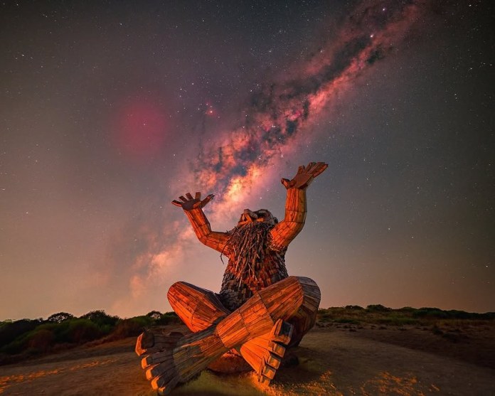 Wooden troll sculpture sitting cross-legged in a desert landscape at night with arms raised toward a vivid Milky Way sky in Mandurah, Western Australia.