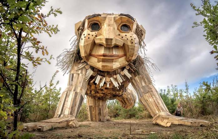 Smiling troll sculpture crawling through a wooded area with braided hair and large hands, surrounded by greenery in Jutland, Denmark.