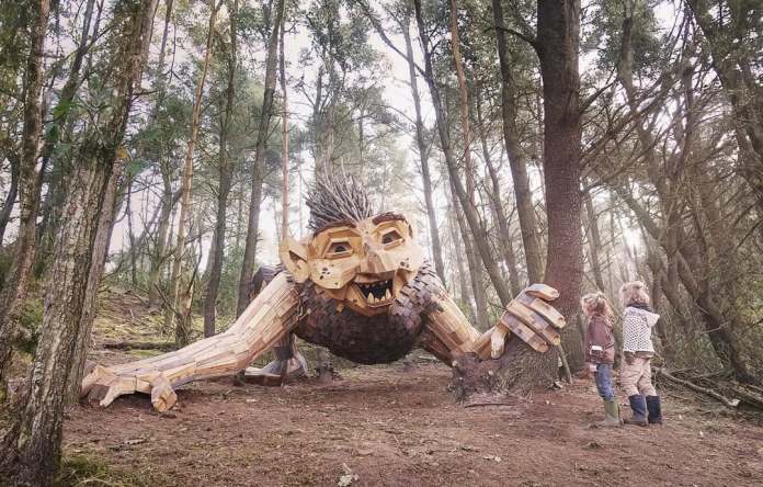 Wooden troll sculpture with wild spiky hair and open mouth, lunging forward and clutching a tree in a forest in Jutland, Denmark.