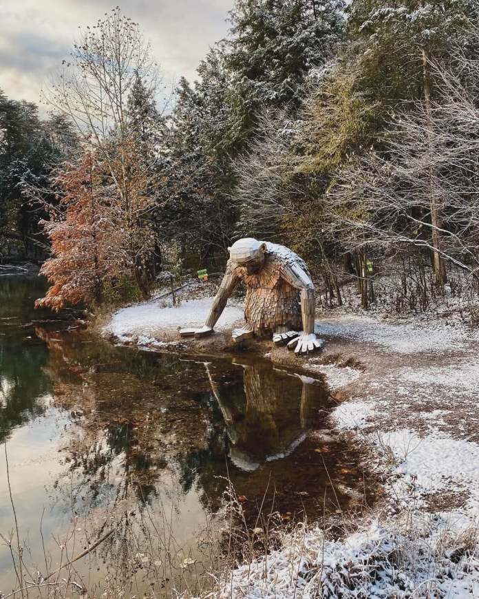 Large wooden troll kneeling beside a reflective lake surrounded by snowy trees in Bernheim Forest, Kentucky, USA.