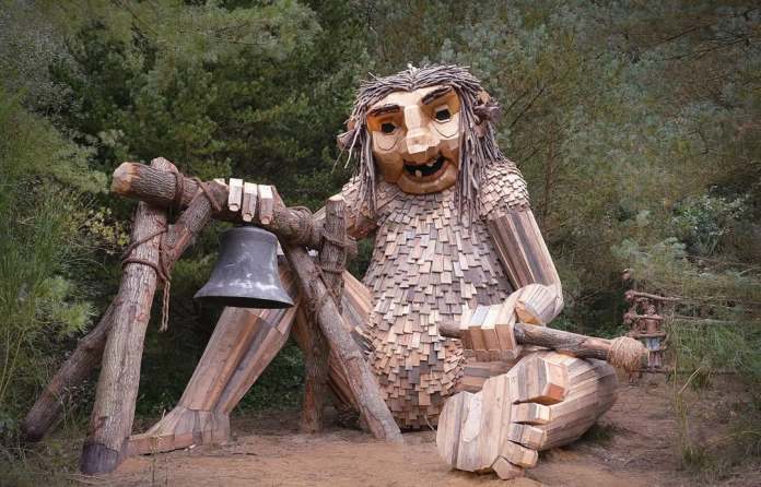 Seated wooden troll sculpture holding a log structure with a large hanging bell, set among pine trees in Jutland, Denmark.