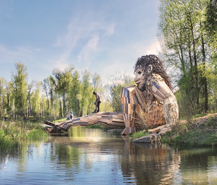 Huge wooden troll sculpture partially submerged in water, forming a bridge with children walking across, located in Rendezvous Park, Wilson, Wyoming.