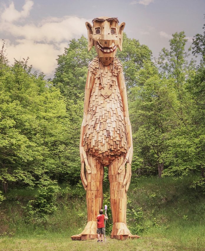 Tallest wooden troll sculpture, standing upright in a forest clearing and smiling down at a small child in red, in Detroit Lakes, Minnesota.