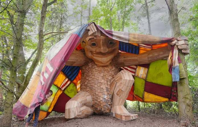 Wooden troll crouching under a bright multicolored fabric tent held up between two trees in a forest in Jutland, Denmark.