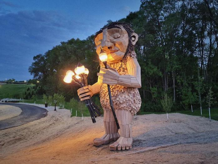 Large wooden troll sculpture by Thomas Dambo named Mama Rosa, standing at dusk on a sandy path holding glowing streetlamps, with forest trees and blue sky in Wauwatosa, USA.
