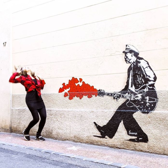 Guitarist mural shooting red hearts from guitar towards a posing pedestrian.