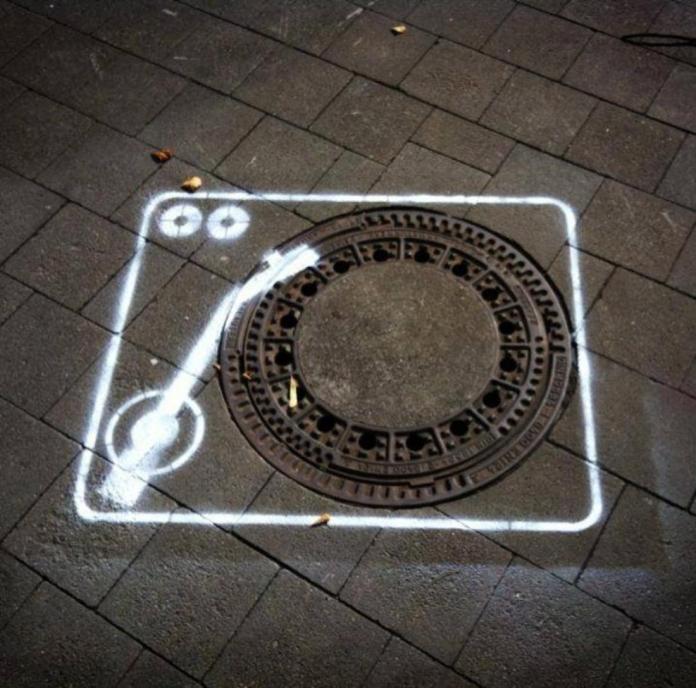 Street art depicting a vinyl record player, using a street manhole as the record and white spray-painted outlines for the player arm and buttons.