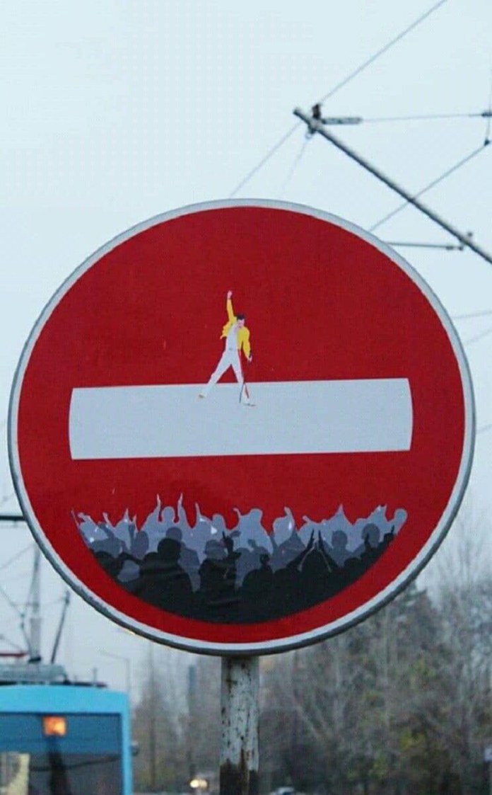 Modified no-entry street sign featuring a small painted figure of Freddie Mercury in his iconic yellow jacket standing on the white bar, with silhouettes of cheering fans below.