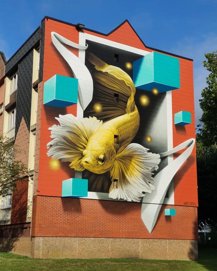 A 3D mural of a gold betta fish appearing to swim out from a hole in a building wall in Abbeville, France, surrounded by turquoise cubes and golden glowing dots.