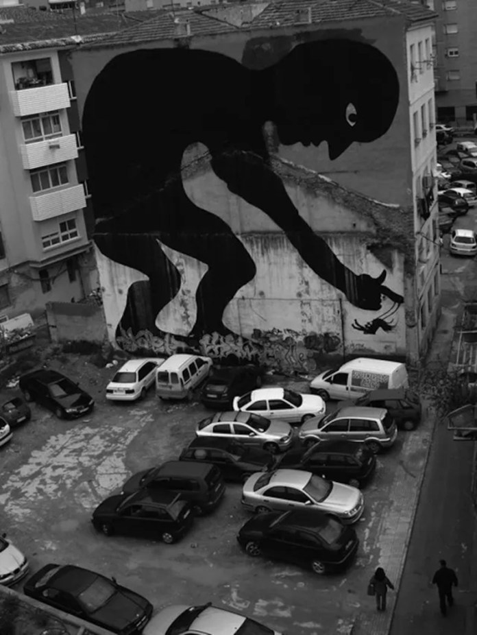 A large black silhouette of a crouched figure painted on a crumbling building wall, reaching toward a person walking through a parking lot in Madrid, Spain.