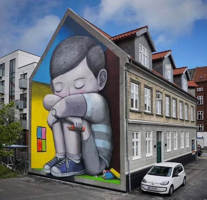 Mural by Seth Globepainter in Aalborg, Denmark, showing a giant boy with closed eyes hugging his knees while holding a crayon, painted on the gable of a house. Background features bright blocks of color and crayons near his feet.