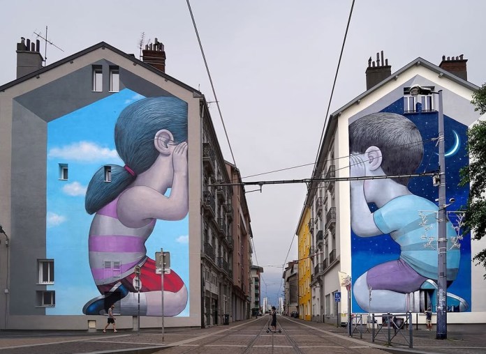 Mural pair by Seth Globepainter in Fontaine, France, with a girl painted on one building whispering toward a boy on another building across the street. One side shows blue sky with clouds, the other a starry night.