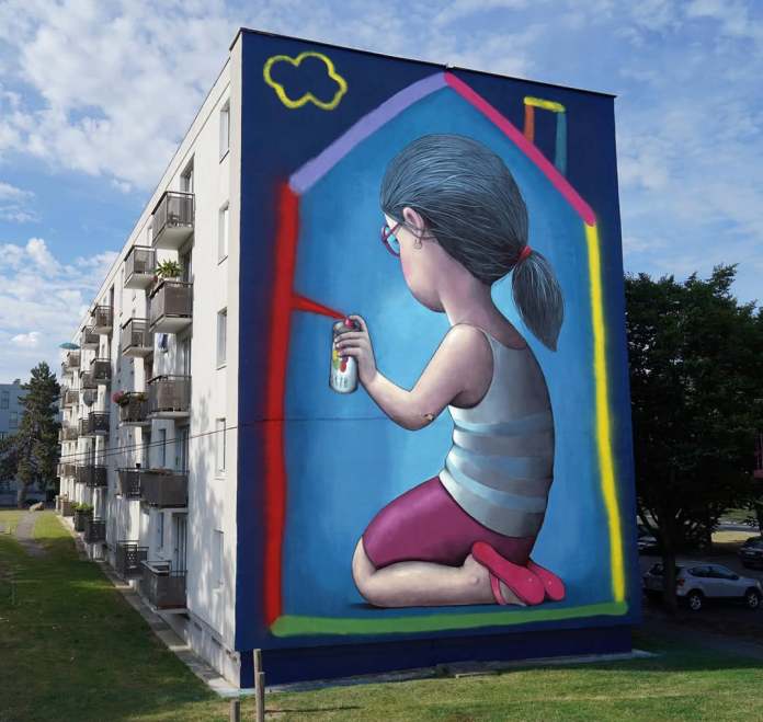 Mural by Seth Globepainter in Laon, France, depicting a girl with glasses kneeling and using a spray can to draw a colorful house outline around herself on a large building wall.