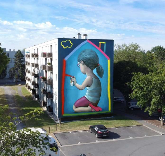 Mural by Seth Globepainter in Laon, France, depicting a girl with glasses kneeling and using a spray can to draw a colorful house outline around herself on a large building wall.