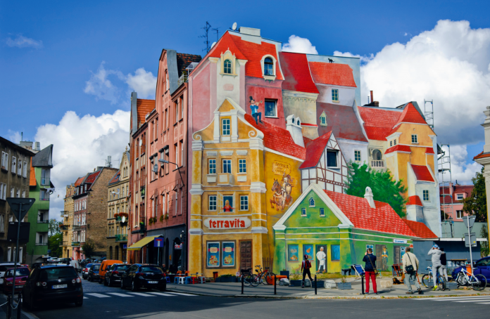A stunning 3D mural by artist Ryszard Paprocki located in the Śródka district of Poznań, Poland. This vibrant artwork transforms the side of a building into a picturesque scene of historic architecture, complete with detailed elements such as a man climbing a ladder, a horse-drawn cart, and lifelike shopfronts. The mural merges seamlessly with its urban surroundings, becoming a beloved landmark that celebrates the area's history. The scene captures a lively street corner filled with pedestrians, bicycles, and cars under a bright blue sky.