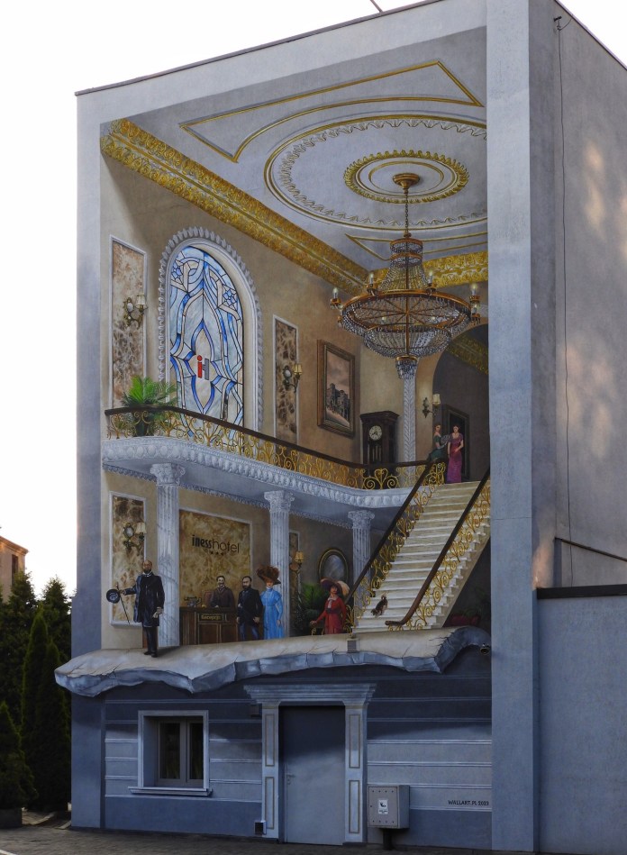Hyper-realistic mural painted on the side of a building in Łódź, Poland, depicting an opulent hotel lobby with chandeliers, gold accents, and guests in period clothing. The illusion includes a detailed staircase and stained-glass windows, simulating interior depth and perspective.