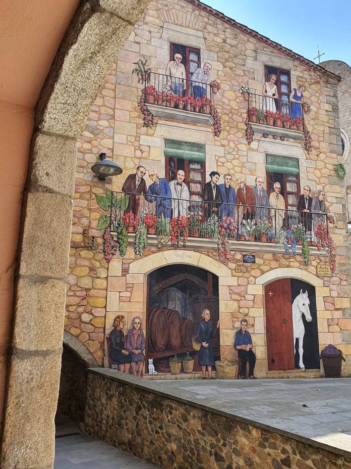 Trompe-l’oeil mural in Calonge, Spain, by Carles Arola, depicting villagers on balconies and at street level with flowers, wine barrels, and a white horse. Painted to appear as part of a stone building facade.