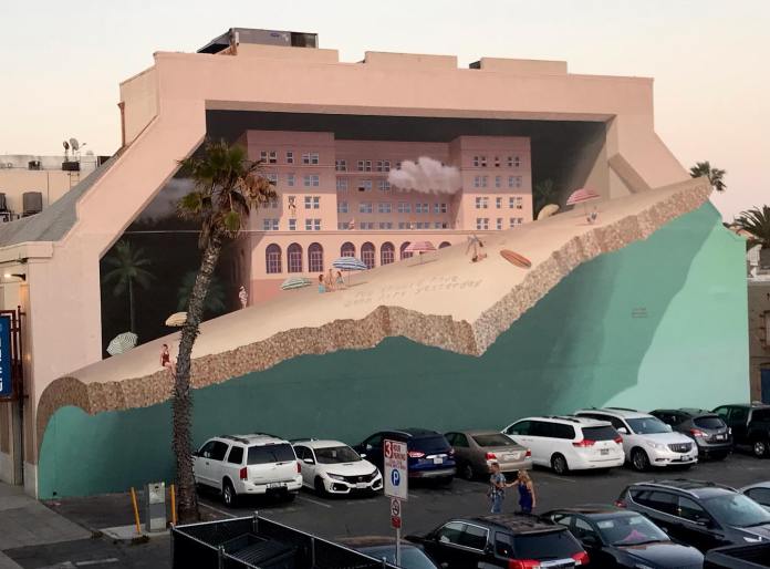 Trompe-l’oeil mural in Hermosa Beach, California, by John Pugh. It depicts a 3D beach with surfers and umbrellas on a steep incline, revealing a pink building behind them. Painted to appear as if the wall has been carved open.