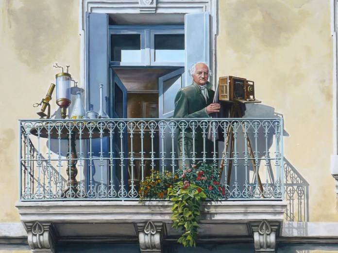 A detailed close-up of Patrick Commecy's mural in Montpellier, France, depicting a distinguished older man with white hair standing on a balcony. He is holding a vintage camera, appearing as a photographer capturing a scene. The balcony railing is ornate, with delicate ironwork, and various scientific instruments, including a telescope and glass beakers, are placed nearby, suggesting an intellectual or artistic pursuit. A lush plant with small red and yellow flowers adds a touch of nature, enhancing the lifelike quality of this trompe-l'œil artwork.