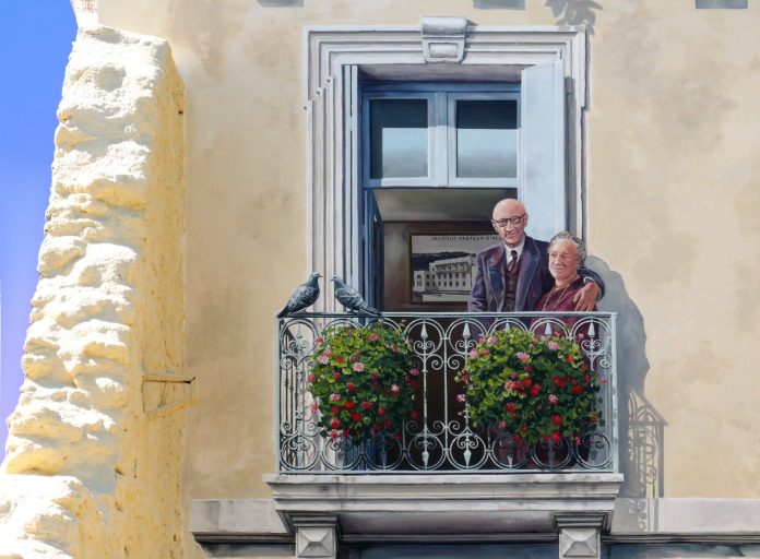 A detailed section of Patrick Commecy's mural in Montpellier, France, showing an elderly couple standing on a balcony, lovingly posed together. The man, wearing glasses and a suit, has his arm around the woman, who is dressed in a maroon outfit with a gentle smile. The balcony railing is adorned with vibrant flower pots, adding a touch of greenery and color to the scene. Two pigeons rest on the railing, enhancing the lifelike feel of the artwork. In the background, a framed illustration of the Institut Pasteur d’Alger can be seen through the open window, referencing historical elements related to the subjects depicted in the mural.