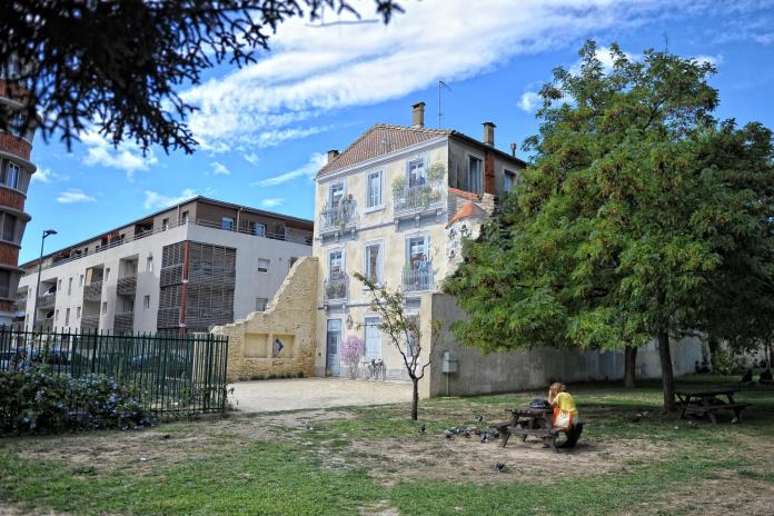 A wide view of a park area in Montpellier, France, showing a striking mural by Patrick Commecy. The mural, painted on the side of a building, features a realistic depiction of a traditional French house with balconies, flowers, and lifelike figures that blend seamlessly into the urban environment. To the right, a large tree provides shade, and a person in a yellow shirt sits on a picnic table in the foreground, surrounded by pigeons. The scene combines modern urban architecture with the charm of classical French artistry, creating a visually engaging juxtaposition between the painted facade and the surrounding park.