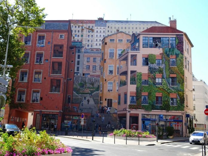 A vibrant view of the 'Wall of the Silk Weavers' mural in Lyon, France, showing the incredible trompe-l'œil artistry that turns the flat building facade into a dynamic urban scene. The mural portrays multiple buildings with various architectural details, including windows, balconies, and staircases that seem to extend into the street. People are painted on the steps, appearing to walk through this bustling neighborhood. Lush greenery covers sections of the wall, blending nature into the scene. Colorful flowerbeds in the foreground add to the charm of this site, complementing the mural’s celebration of Lyon’s historical silk industry.
