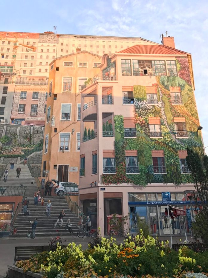 A close-up of the 'Wall of the Silk Weavers' mural in Lyon, France, capturing the intricate details that make this trompe-l'œil artwork so lifelike. The mural features painted buildings with balconies, windows, and a staircase filled with people engaged in everyday activities, creating the illusion of a bustling neighborhood. The right side is adorned with lush greenery that appears to grow up the wall, blending with the architectural elements. The scene is illuminated by soft sunlight, enhancing the vibrant colors and textures. A bed of colorful flowers in the foreground adds a real touch of nature to the painted urban landscape.