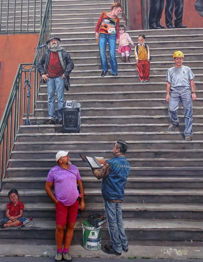 A close-up of the 'Wall of the Silk Weavers' mural in Lyon, France, showing the realistic painted figures on the staircase. The mural captures a variety of people in different poses, including a man with a microphone and speaker as if giving a speech or performing, a mother guiding her young daughter down the steps, a worker in a hard hat, and a painter with a palette and brush. Each figure is painted with detailed expressions and attire, blending seamlessly into the stairwell scene. A man in a bright purple shirt and red shorts, who is standing in front of the mural, looks up at the painted artist as if interacting with him, adding a playful, immersive element to the artwork.