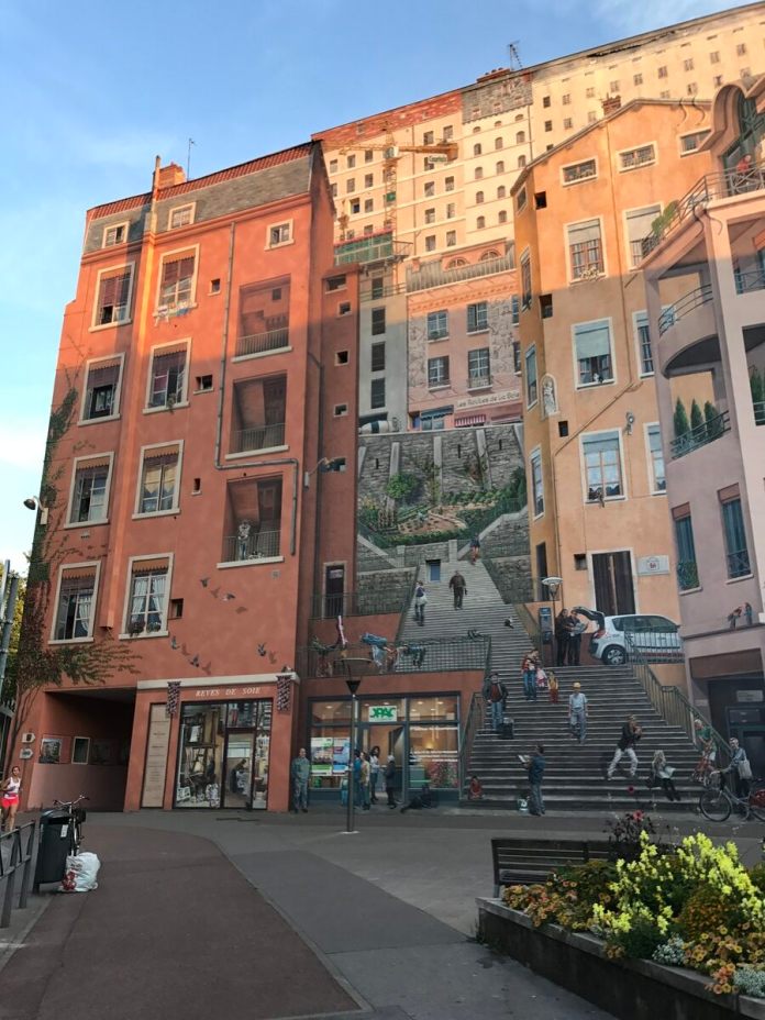 A wide-angle view of the 'Wall of the Silk Weavers' mural in Lyon, France, capturing the detailed trompe-l'œil artistry that transforms the building facade into a lively urban scene. The mural spans multiple stories, depicting buildings with balconies, windows, and a central staircase filled with painted figures going about their day. Shops and storefronts at the ground level appear three-dimensional, blending seamlessly with the real street environment. Flower beds in the foreground add color, complementing the warm hues of the mural under soft evening light. The mural pays homage to Lyon’s rich history in the silk weaving industry, bringing vibrant life and charm to this urban space.