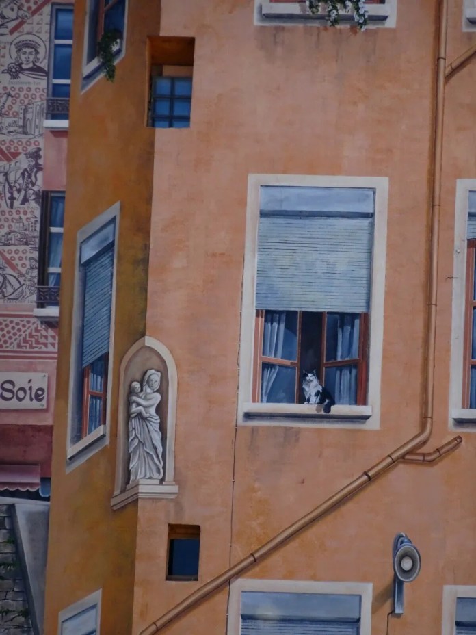 A close-up section of the 'Wall of the Silk Weavers' mural in Lyon, France, showcasing the remarkable attention to detail in this trompe-l'œil artwork. The focus is on a corner of a painted building, where a small sculpted figure in a niche adds an architectural touch. Below it, a realistic painted window reveals a black and white cat sitting on the windowsill, gazing out as if observing the street below. The colors and shading create a lifelike effect, blending seamlessly with the surrounding painted textures, pipes, and window shutters, enhancing the mural's illusion of depth and realism.
