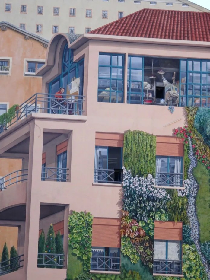 A detailed view of the 'Wall of the Silk Weavers' mural in Lyon, France, focusing on the upper floors of a painted building. The artwork features a balcony where an artist in red is depicted painting at an easel, adding a meta element to the mural. Through the large, glass-paneled windows, various objects, including lights and furniture, can be seen, creating a realistic interior scene. The building facade is adorned with painted greenery, with lush vines and flowering plants climbing the walls, adding a natural element that complements the architectural details. The red-tiled roof and soft pastel colors bring warmth to the scene, enhancing the mural's lifelike illusion.