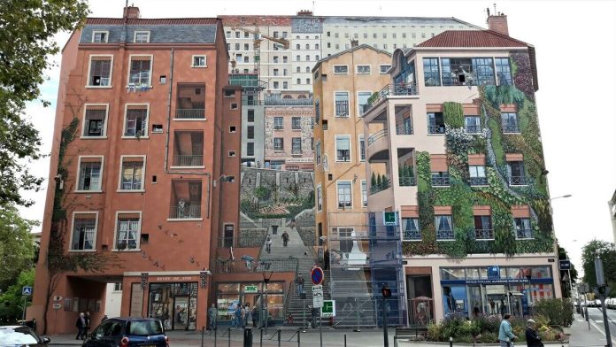 A close-up view of the 'Wall of the Silk Weavers' mural in Lyon, France, capturing the stunning trompe-l'œil effect that transforms a flat wall into a vibrant cityscape. The mural displays a realistic depiction of French architecture, with windows, balconies, and staircases that seem three-dimensional. People appear to walk up the stairs and interact in this imagined urban environment, giving the illusion of a bustling neighborhood scene. The right side of the mural is covered in painted greenery, mimicking ivy and other plants growing up the facade. The artwork beautifully integrates elements of daily life, architecture, and nature, honoring Lyon’s heritage in the silk weaving industry.