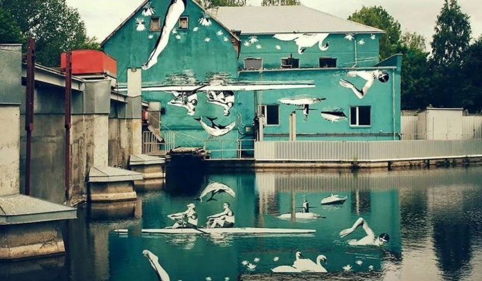 A turquoise mural by artist Ray Bartkus in Marijampolė, Lithuania, painted on the side of a building by a canal. The mural depicts an upside-down scene of swimmers, rowers, and swans, which cleverly reveals its full meaning when reflected in the water, creating an upright and seamless composition of people swimming, rowing, and swans floating on a serene lake surrounded by lily pads.