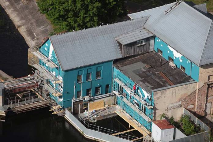 An aerial view of the turquoise mural by artist Ray Bartkus in Marijampolė, Lithuania, painted on the side of a building adjacent to a canal. The image shows the mural in progress, with scaffolding and workers visible as they paint the upside-down design. The mural is designed to reveal its full meaning when reflected in the water, creating an upright scene of swimmers, rowers, and swans.