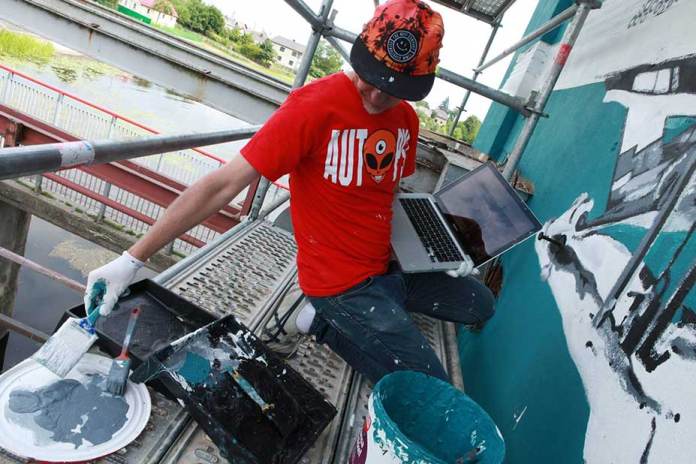 Artist Ray Bartkus working on his upside-down mural in Marijampolė, Lithuania. He is sitting on scaffolding, holding a laptop in one hand and painting tools in the other, with a bucket of turquoise paint and brushes nearby. The mural is in progress on the building wall, featuring intricate details that transform into a cohesive scene when reflected in the canal below.