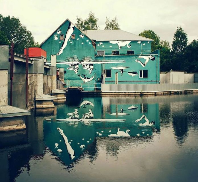 A turquoise mural by artist Ray Bartkus in Marijampolė, Lithuania, painted on the side of a building by a canal. The mural depicts an upside-down scene of swimmers, rowers, and swans, which cleverly reveals its full meaning when reflected in the water, creating an upright and seamless composition of people swimming, rowing, and swans floating on a serene lake surrounded by lily pads.