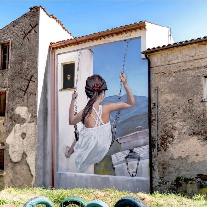 Mural of a girl in a white summer dress swinging out from a building facade as if suspended in air, painted with photorealistic detail against a backdrop of Italian rooftops and distant blue mountains.