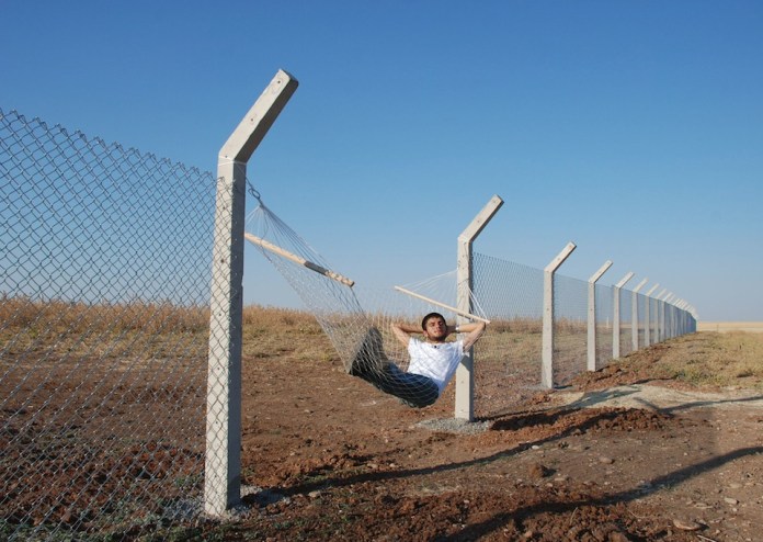 Installation artwork showing a man lying in a hammock made from cut metal fencing, suspended between angled concrete border posts in a barren field.