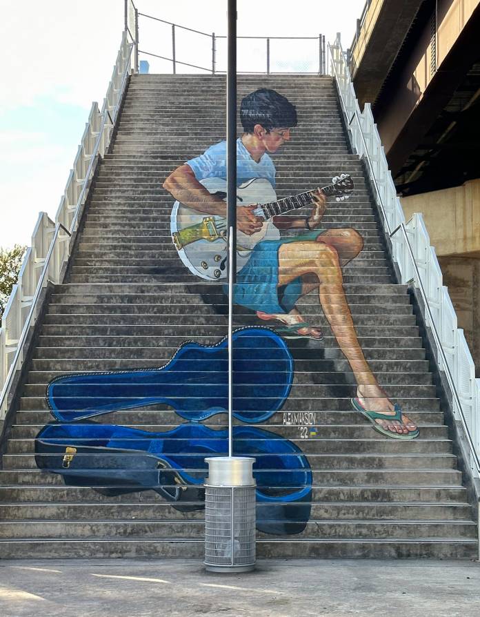 Mural painted across concrete stairs of a boy in flip-flops playing guitar, with a realistic blue guitar case resting at the base of the steps.