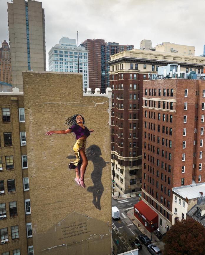 Mural on high-rise building showing a woman in a burgundy top and yellow pants jumping upward, casting a shadow onto the wall with city buildings in background.