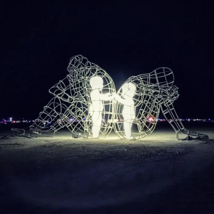Wireframe sculptures of two adults sitting back-to-back, each containing a glowing child figure inside reaching toward the other, lit up at night on a desert plain.