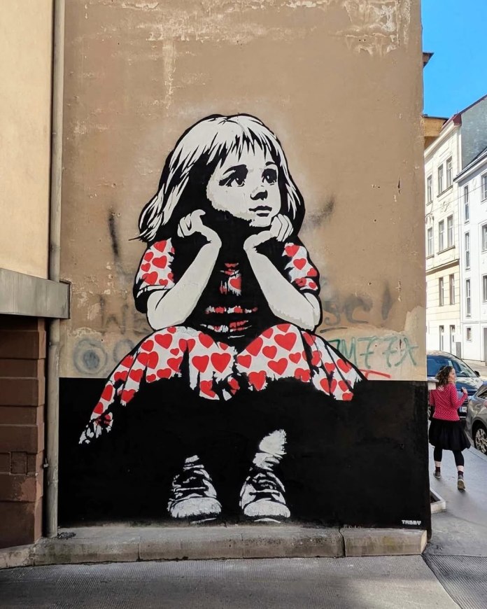 Black-and-white stencil artwork of a young girl in red dress with heart prints, sitting on a black base with chin resting on hands, painted by TABBY on a corner wall in Vienna.