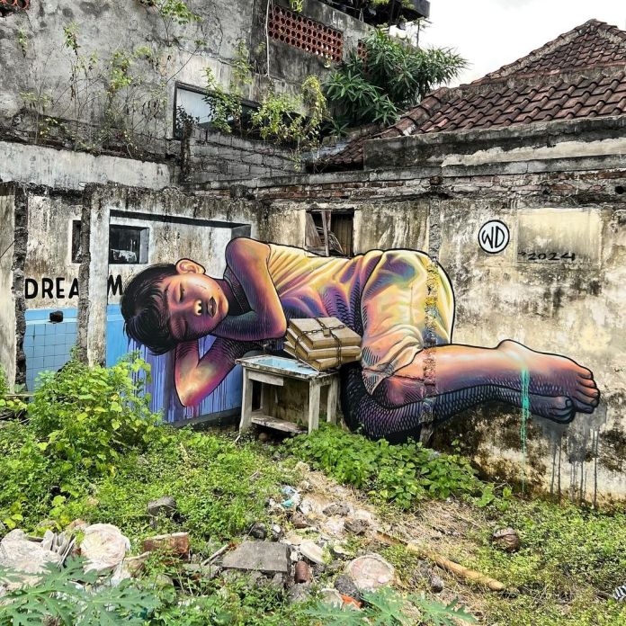 Mural of a boy with colorful shading sleeping curled up on an abandoned building, partially interacting with walls and debris in Denpasar, Bali.