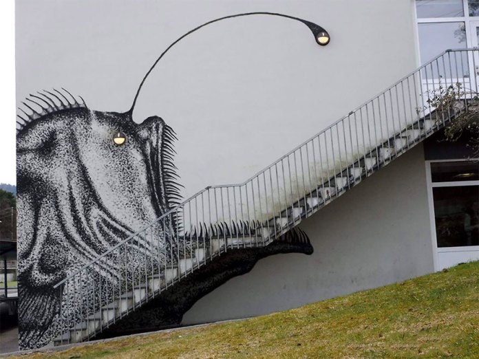 A striking street art mural by SKURK at Rå Skole in Bergen, Norway, creatively incorporates architectural features into its design. The mural depicts an anglerfish, with its body painted along the side of a white building, and a staircase cleverly forming the fish's mouth and teeth. A streetlight at the top of the staircase doubles as the anglerfish's glowing lure. The artwork is equally impactful during the day, showcasing its detailed black-and-white stippling technique, and at night, when the illuminated light transforms the mural into a vivid and dynamic scene. This masterpiece exemplifies the seamless blending of art and architecture.