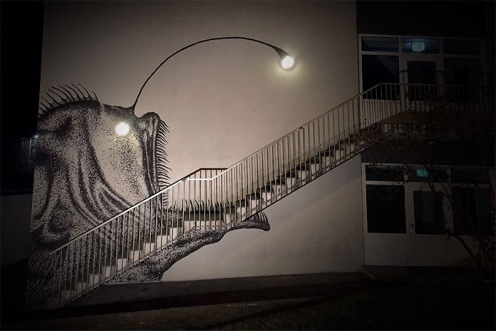 A striking street art mural by SKURK at Rå Skole in Bergen, Norway, creatively incorporates architectural features into its design. The mural depicts an anglerfish, with its body painted along the side of a white building, and a staircase cleverly forming the fish's mouth and teeth. A streetlight at the top of the staircase doubles as the anglerfish's glowing lure. The artwork is equally impactful during the day, showcasing its detailed black-and-white stippling technique, and at night, when the illuminated light transforms the mural into a vivid and dynamic scene. This masterpiece exemplifies the seamless blending of art and architecture.