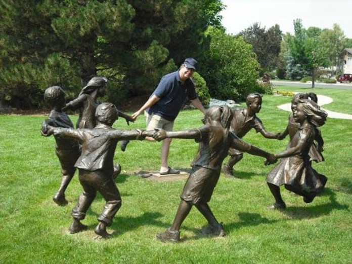 A man in a navy shirt and cap joins hands with bronze children in a ring-shaped sculpture on a grassy park lawn, appearing to be part of their playful game.