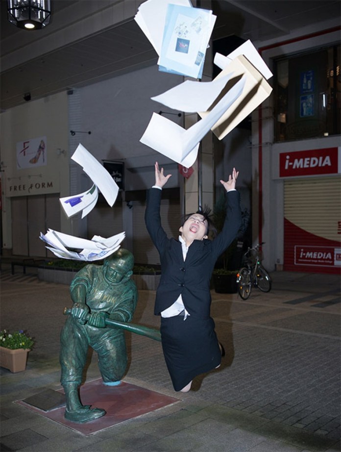 A statue of a boy swinging a bat appears to have hit a woman midair as she jumps, arms up, with papers flying above her in a Japanese shopping arcade.