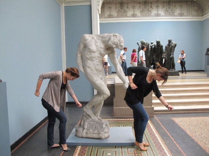 Two women in a museum lean forward with arms bent beside a white marble sculpture doing the same pose, visually merging into the figure’s movement.